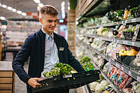 Ein junger Mensch steht vor einem Gemüseregal im Supermarkt und trägt einen Korb mit Gemüse zum Verräumen