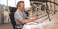 Ein Mann mit Headset sitzt in einer Leitstelle an einem großen Bedienpult mit mehreren Monitoren, Telefonen und Mikrofonen. Er lächelt und arbeitet konzentriert an der Steuer- oder Kommunikationsanlage, während im Hintergrund weitere Arbeitsplätze zu sehen sind.