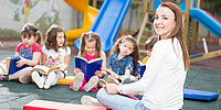Foto einer Erzieherin sowie Kinder, die lesen. Im Hintergrund ist eine Rutsche zu sehen.