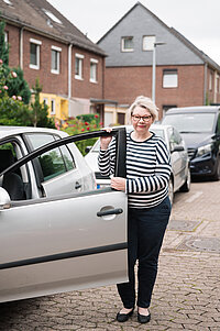 Eine Frau steht neben einem geparkten Auto und hält die geöffnete Fahrertür fest. Die Aufnahme entstand in einer Wohnstraße mit Reihenhäusern und Gärten im Hintergrund.
