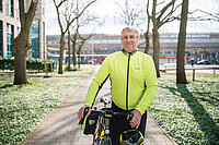 Mark Tchernine steht in heller Fahrradkleidung mit seinem Fahrrad auf einem Weg in einer Grünanlage und blickt in die Kamera. Er hält einen Fahrradhelm in der Hand.