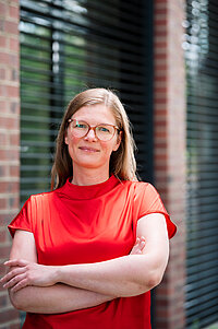 Eine Frau mit langen hellbraunen Haaren und Brille steht mit verschränkten Armen vor einer Backsteinfassade. Sie trägt eine leuchtend rote Bluse und lächelt in die Kamera.