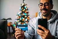 Ein Mann schaut vor einem Weihnachtsbaum auf seine Bankkarte und lacht Ein Mann schaut vor einem Weihnachtsbaum auf seine Bankkarte und lacht
