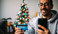 Ein Mann schaut vor einem Weihnachtsbaum auf seine Bankkarte und lacht