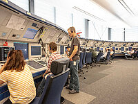 Fluglotsinnen und Fluglotsen sitzen in der Radarkontrollzentrale Bremen an ihren Arbeitsplätzen, Sven Wilke steht zwischen ihnen und spricht mit einer Kollegin.