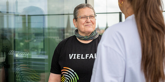 Person mit schwarzem T-Shirt auf dem Vielfalt steht spricht mit einer anderen Person.