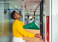 Eine Frau steht mit einer Kiste mit Büromaterial im Flur und schließt die Augen