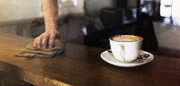 Person wischt eine Theke ab auf der eine Tasse Kaffee steht.