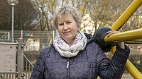 Eine Frau mit kurzer grauer Frisur steht auf einem Spielplatz. Sie trägt eine dunkle Jacke und einen hellen Schal und hält sich an einem gelben Klettergerüst fest. Im Hintergrund stehen kahle Bäume und weitere Spielgeräte.