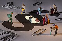 Diverse Miniaturfiguren, die ihrer Arbeit nachgehen - auf dem Boden liegen Büroklammern sowie ein Großes § Zeichen.