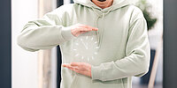 Eine Person im hellgrünen Hoodie hält die Hände waagerecht übereinander, als würde sie einen Zeitraum anzeigen. Zwischen den Händen ist grafisch eine Uhr eingeblendet, die Zeit oder Arbeitszeit symbolisiert.