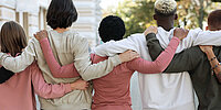 Fünf junge Menschen stehen nebeneinander im Freien und haben die Arme freundschaftlich umeinander gelegt. Sie sind von hinten zu sehen und wirken solidarisch und verbunden.