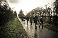 Eine Gruppe von Menschen wandert an einem grauen Wintertag über einen nassen Weg durch eine ländliche Landschaft. Eine Person zieht einen Bollerwagen, im Hintergrund steht ein Windrad.