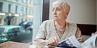 Ältere Frau sitzt in einem Café, liest Zeitung und blickt nach draußen.