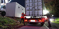 Zwei Arbeiter verladen bei Dunkelheit einen großen Container auf einen Lkw. Einer trägt eine Warnweste, der andere steht seitlich am Fahrzeug; Scheinwerfer beleuchten die Szene, im Hintergrund ist weiteres Gerät zu sehe