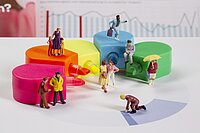 Diverse Miniaturfiguren, die vor bunten Textmarkern stehen. Im Hintergrund ist ein Graph zu sehen.