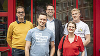 Vier Männer und eine Frau stehen lächelnd vor dem roten Eingang der Arbeitnehmerkammer Bremen. Ein Mann trägt ein T-Shirt mit der Aufschrift „Für sichere Häfen! An der Küste und überall in Europa“. Zu sehen sind Ralf Wilke, Felix Gröll, Christian Wechselbaum, Daphne Weber und Nils Wolpmann (von links nach rechts).