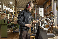 Eine Person arbeitet in einer Holzwerkstatt an einer Drechselmaschine. Sie trägt Gehörschutz und Schutzbrille und bearbeitet ein eingespanntes Holzstück, während Holzspäne umherfliegen. Im Hintergrund sind weitere Maschinen, Werkzeuge und Arbeitsmaterialien zu sehen.