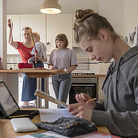 Jugendlich am Schreibtisch, im Hintergrund eine Mutter mit Kind auf dem Arm, die in der Küche mit einer weiteren Jugendlichen steht.