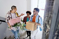 Drei junge Menschen stehen in einem hellen Treppenhaus und tragen Kartons und Kisten für einen Umzug
