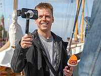 Nils Fricke, Volontär bei Radio Bremen, filmt mit einem Smartphone auf einem Gimbal und hält ein orangefarbenes Mikrofon in der Hand.