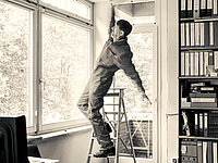 Schwarz-Weiß-Fotografie eines Büroraums mit großen Fenstern und einem Regal voller Aktenordner. Ein Mann steht auf einer Klappleiter und streckt sich weit nach oben, um einen Bereich über einem Fenster zu erreichen. Dabei gerät er sichtbar aus dem Gleichgewicht und lehnt sich nach hinten, sodass eine Sturzgefahr entsteht. Im Hintergrund sind Fensterfronten und Bäume außerhalb des Gebäudes zu sehen.