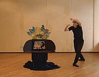 Eine in schwarzgekleidete Person performt in Richtung eines Puppentheater ähnlichen Aufbaus.