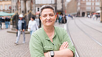 Eine Frau mit kurzen grauen Haaren und grünem Hemd steht mit verschränkten Armen auf dem Bremer Marktplatz. Im Hintergrund sind der Roland, historische Gebäude und Straßenbahnschienen zu sehen.