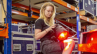 Eine Veranstaltungstechnikerin prüft in einem Lager eine rot leuchtende Bühnenlampe. Um sie herum stehen Regale mit großen Transportkisten und weiterem Bühnenequipment.