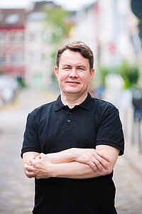 Ein Mann mit kurzen braunen Haaren und schwarzem Poloshirt steht mit verschränkten Armen in einer Straße. Im Hintergrund sind unscharf Häuser und Bäume zu sehen.
