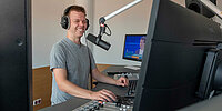 Mann in einem Studio bzw. vor einem Mischpult. Er trägt Kopfhörer und spricht in ein Mikrofon.