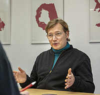 Peer Rosenthal gestikuliert im Interview am Tisch sitzend, im Hintergrund Kunst an der Wand