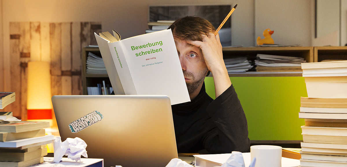 Mann am Schreibtisch auf dem mehrere Dokumente sich stapeln, fasst sich fragend an den Kopf und liest ein Buch.